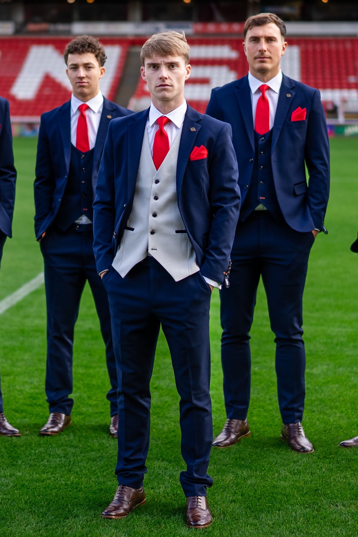 Barnsley FC captain wearing the SUITBAE Charles Navy Contrast Three-Piece Suit with a light grey waistcoat, red tie, and matching pocket square, styled with brown brogues. He stands confidently on the pitch with teammates in matching navy suits behind him.