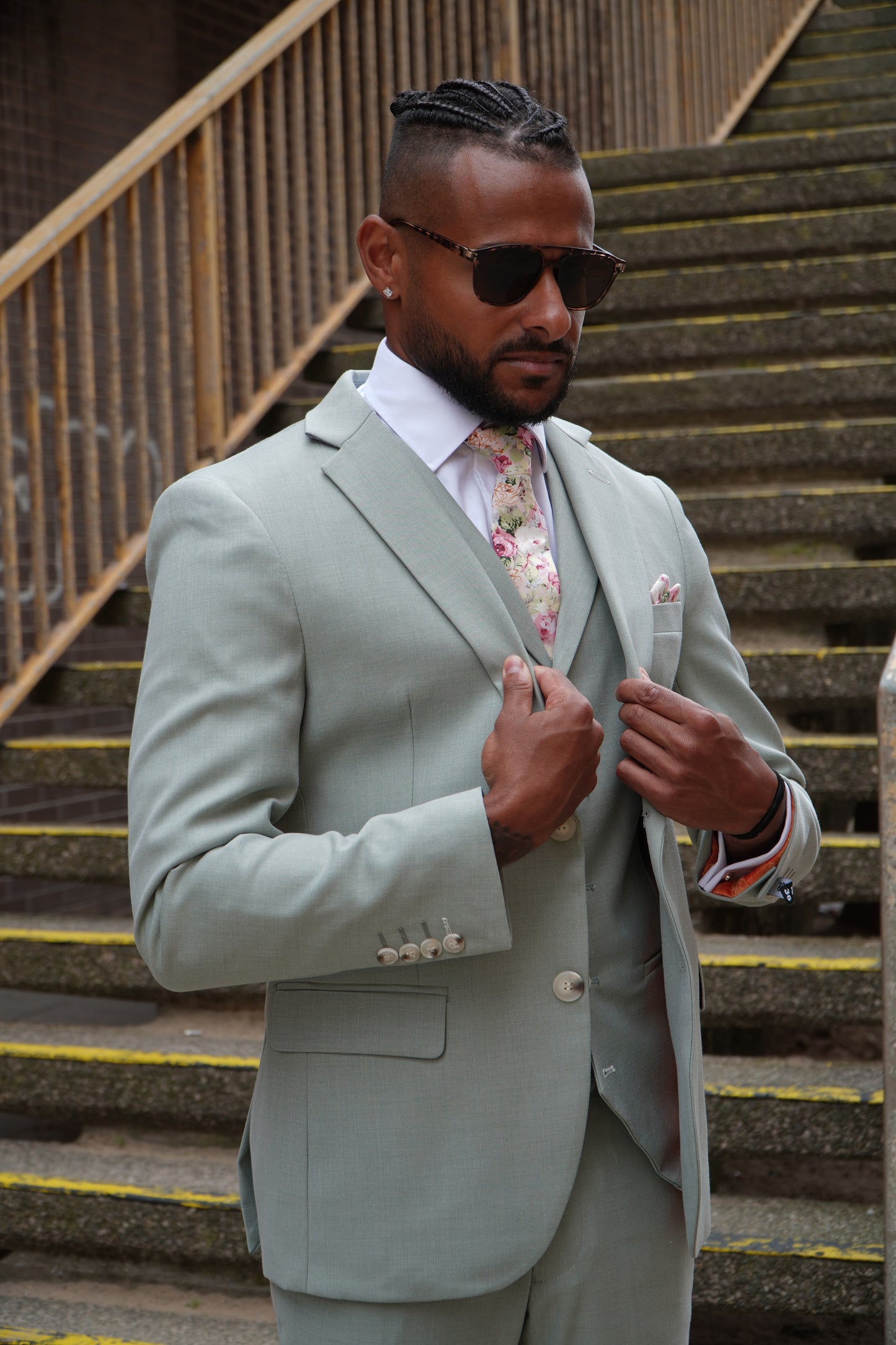 Model wearing the SUITBAE Callum Sage Three-Piece Suit — tailored light sage green fabric with matching waistcoat and trousers, styled with a floral tie and pocket square for weddings and formal summer occasions.