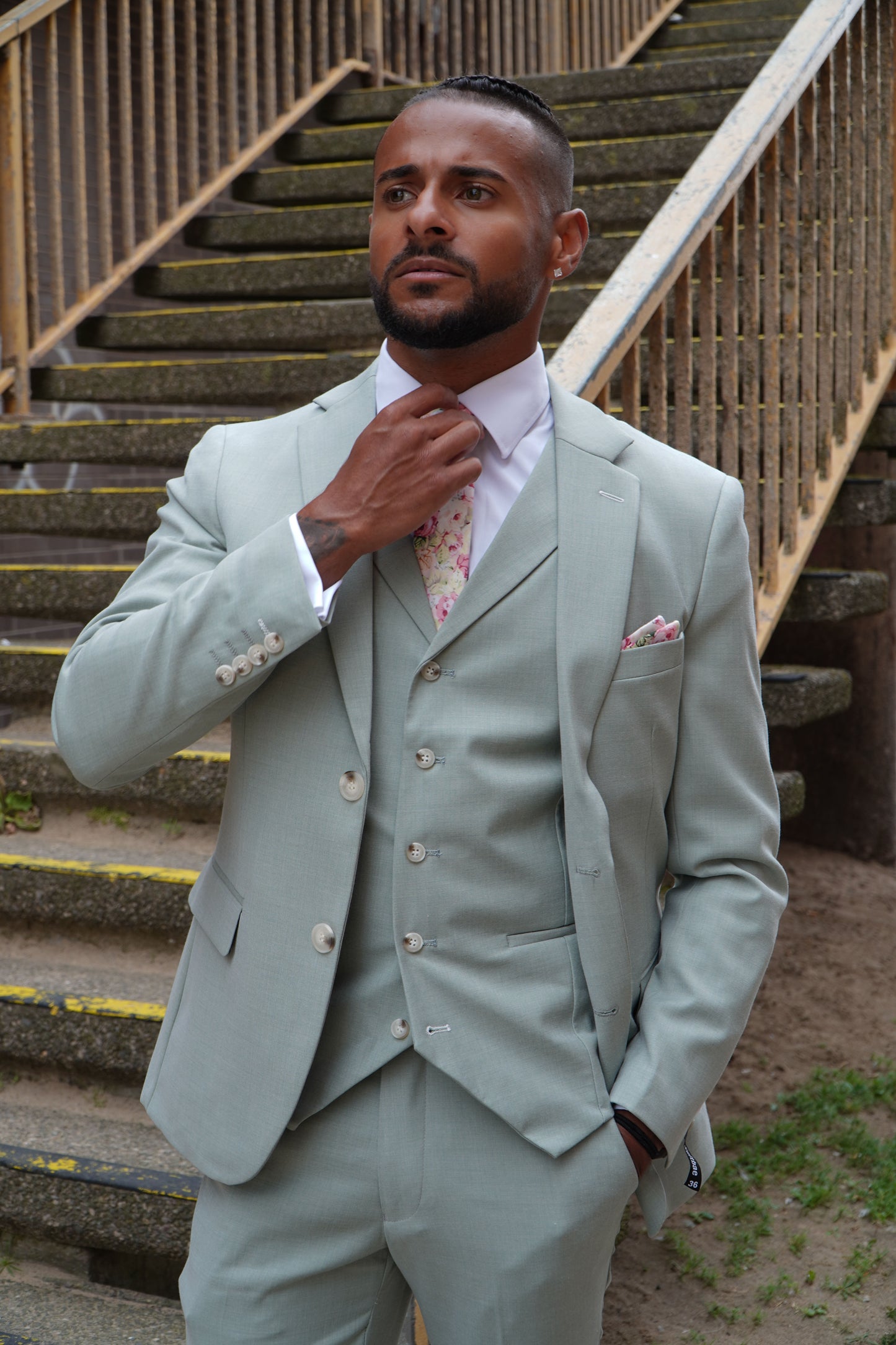 Model wearing the SUITBAE Callum Sage Three-Piece Suit — tailored light sage green design with matching waistcoat and trousers, styled with a floral tie and pocket square for weddings and summer occasions.