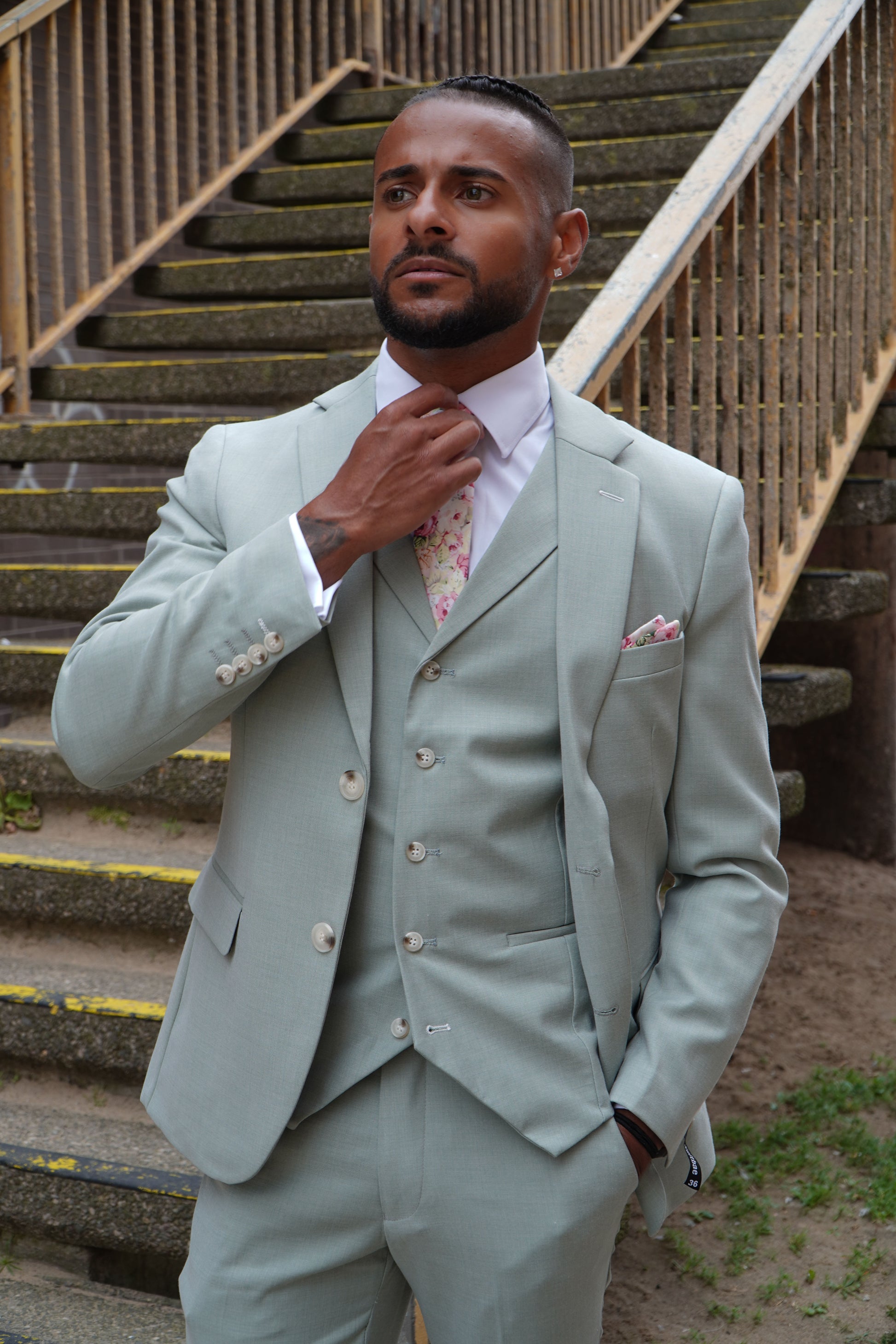 Model wearing the SUITBAE Callum Sage Three-Piece Suit — tailored light sage green design with matching waistcoat and trousers, styled with a floral tie and pocket square for weddings and summer occasions.