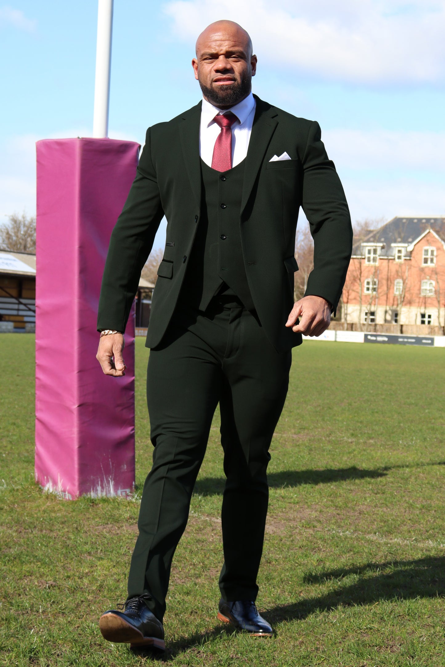 Model wearing the SUITBAE Charles Green Three-Piece Suit on a rugby field. The suit features a structured green blazer, horseshoe-cut waistcoat, and slim-fit trousers, styled with a white shirt and burgundy tie — ideal for athletic or plus-size builds seeking a sharp, confident fit.