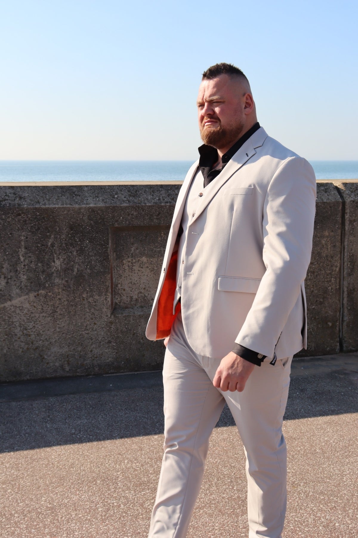 Full view of the SUITBAE Kian Stone Three-Piece Suit — stone beige with contrast brown buttons and orange inner lining. Shown with a black shirt for a bold formal look. Available in plus sizes up to 58" chest.
