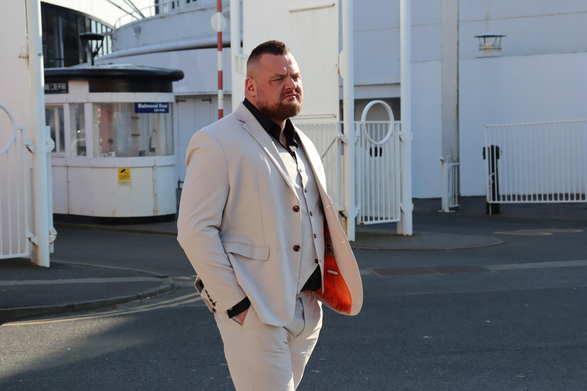 Front view of the SUITBAE Kian Stone Three-Piece Suit — tailored in stone beige with brown buttons, orange inner lining, and a black shirt contrast. Available in plus sizes for weddings, races, and formal summer wear.