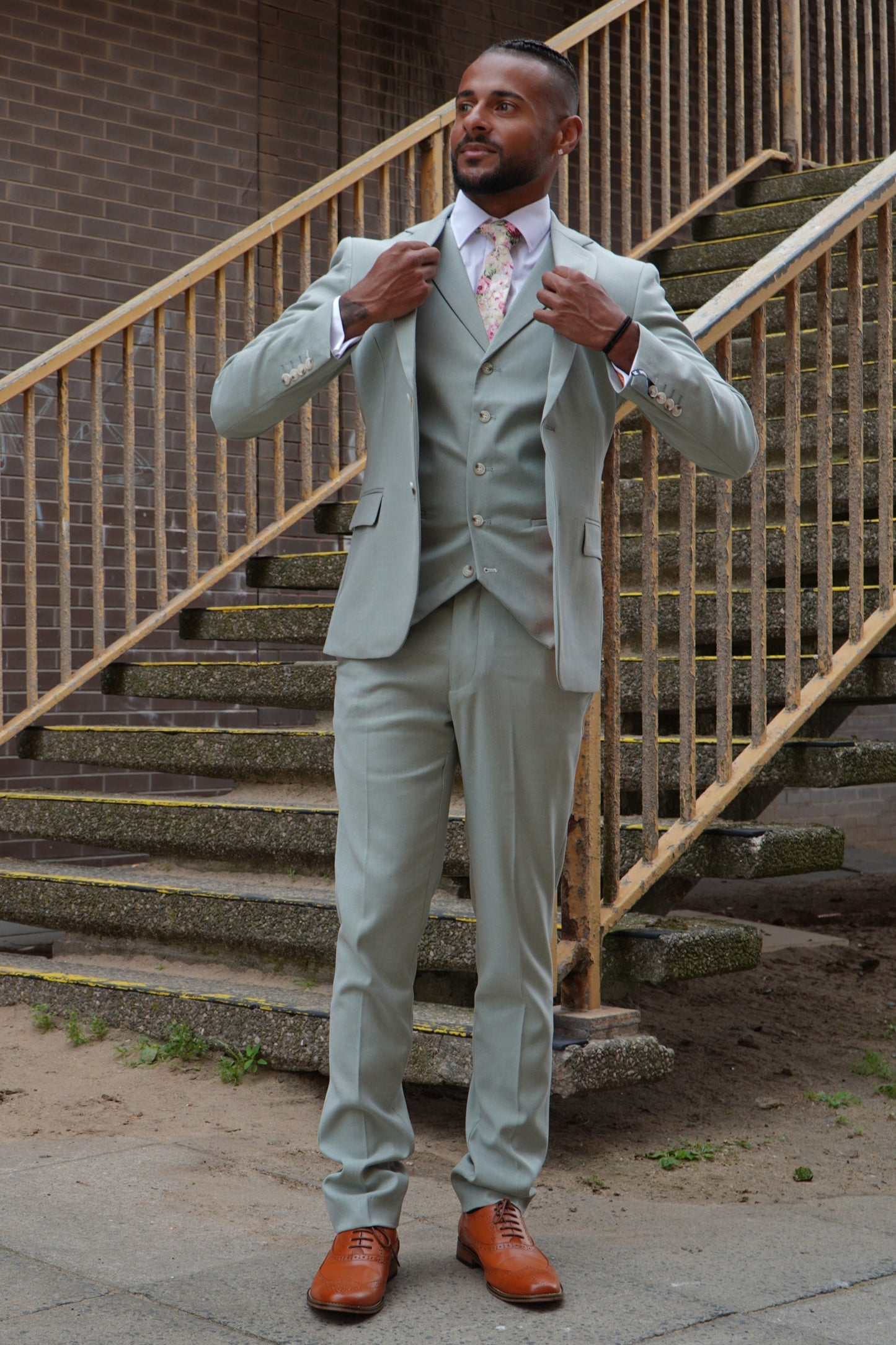 Model wearing the SUITBAE Callum Sage Three-Piece Suit — light sage green slim-fit design with matching waistcoat and trousers, styled with a floral tie and tan brogues for weddings and summer events.