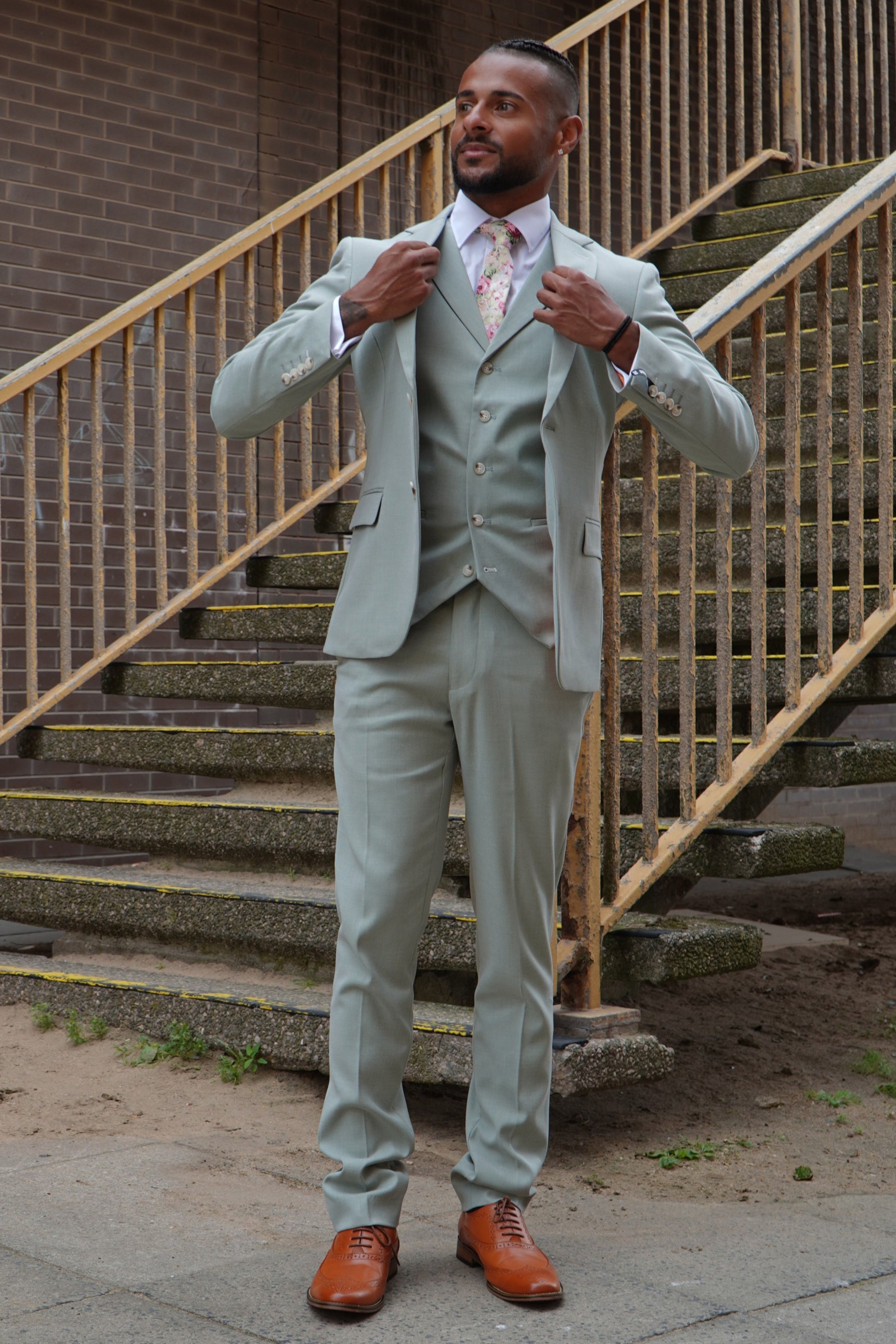 Model wearing the SUITBAE Callum Sage Three-Piece Suit — light sage green slim-fit design with matching waistcoat and trousers, styled with a floral tie and tan brogues for weddings and summer events.