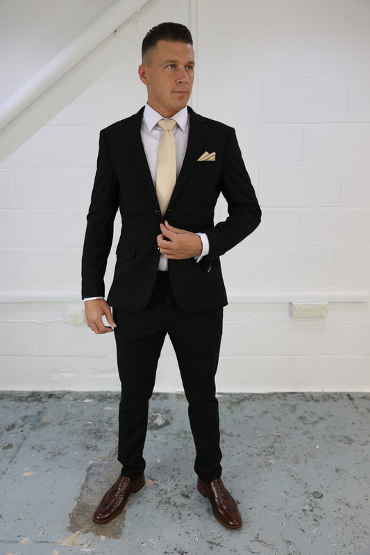 Man wearing a Suitbae black pinstripe two-piece suit, featuring a tailored blazer and matching slim-fit tapered trousers, styled with a white shirt, gold tie, and pocket square; ideal for formal events or smart-casual wear.