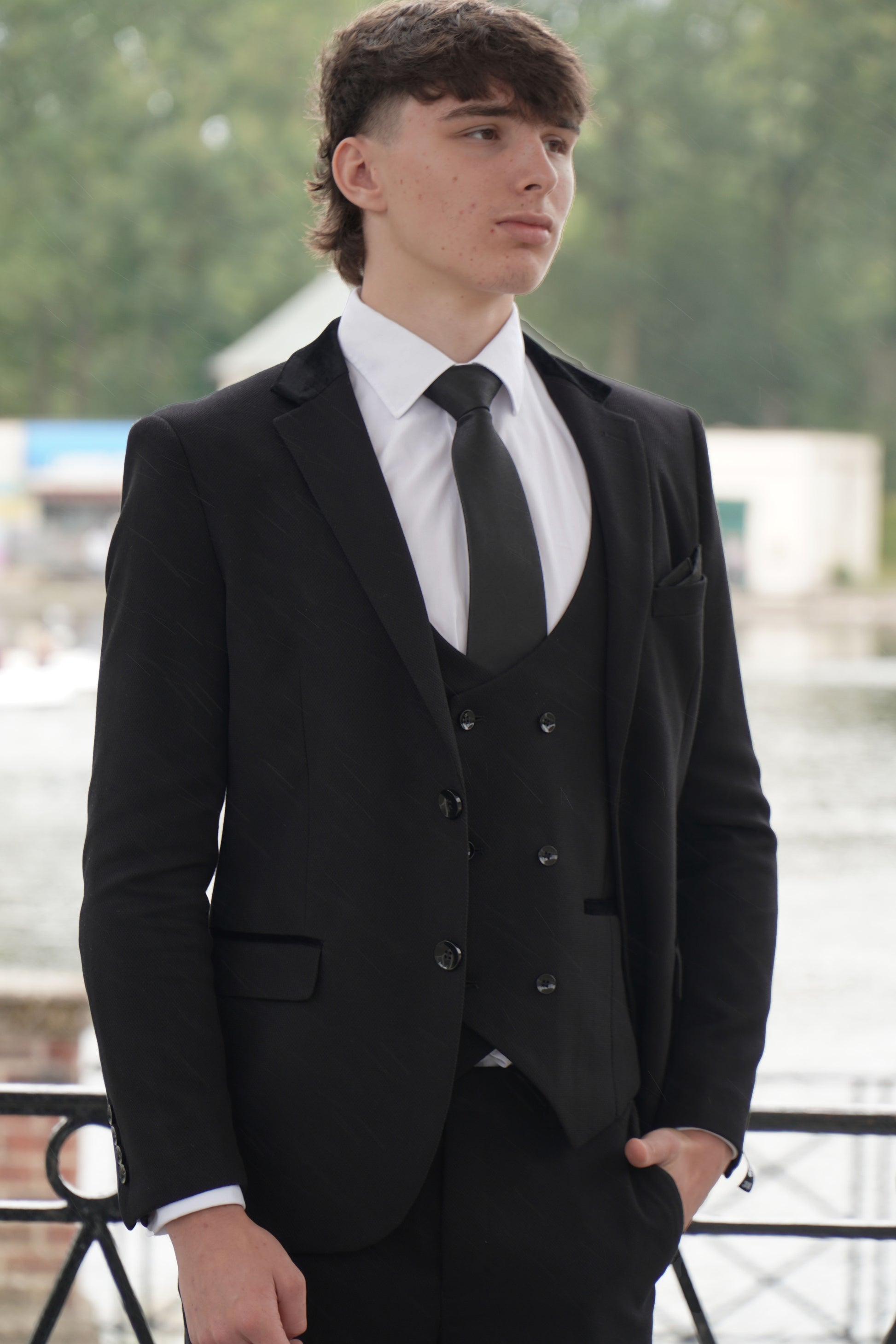 Model wearing the SUITBAE Majid Textured Three-Piece Suit — sleek black tailored design with subtle fabric texture, styled with a white shirt and black tie for weddings, proms and formal occasions.