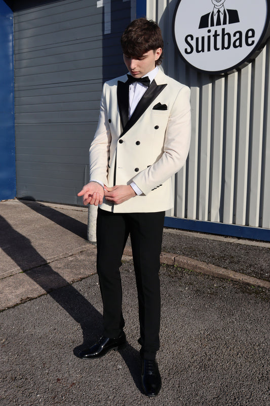 Model standing outside the SUITBAE showroom adjusting his cuff while wearing the SUITBAE Ivory Double-Breasted Tuxedo, featuring black satin peak lapels, contrast buttons, and pocket trim, paired with black trousers, bow tie and patent black shoes.