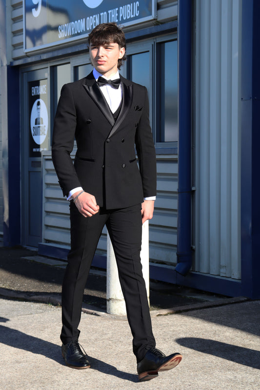 Model walking outside the SUITBAE showroom wearing the SUITBAE Diablo Black Double-Breasted Tuxedo with satin peak lapels, black buttons, and matching trousers, styled with a white shirt, black bow tie, and black formal shoes.