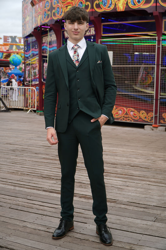 Model wearing the SUITBAE Archie Green Three-Piece Suit — dark green slim-fit design with matching waistcoat and trousers, styled with a floral tie for weddings, proms and formal occasions.