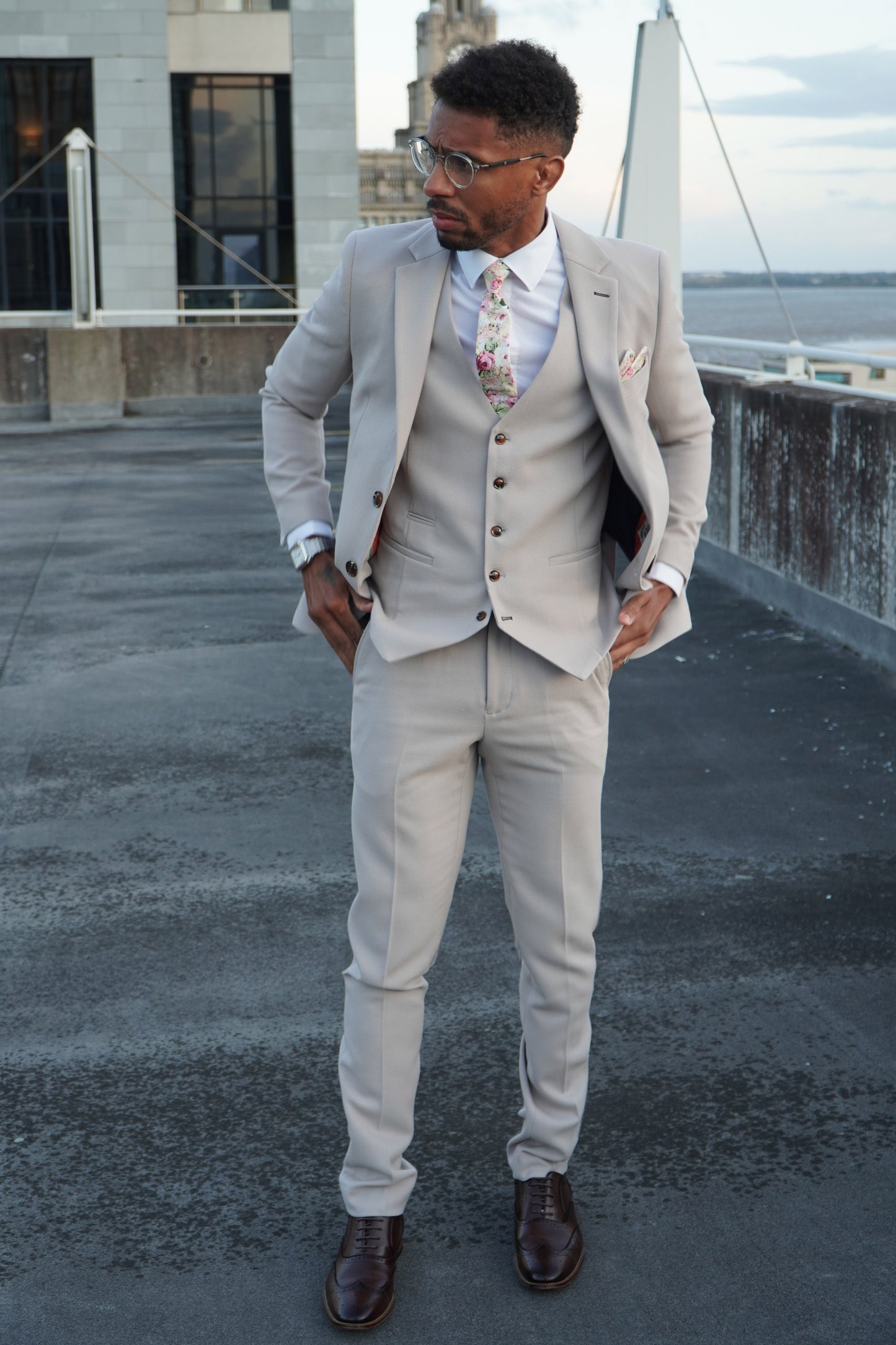 Model wearing the SUITBAE Escobar Stone Three-Piece Suit — slim-fit beige design with brown buttons, paired with a floral tie and dark brown shoes, ideal for weddings and summer formal occasions.