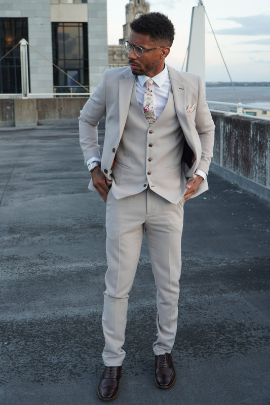 Model wearing the SUITBAE Escobar Stone Three-Piece Suit — slim-fit beige design with brown buttons, paired with a floral tie and dark brown shoes, ideal for weddings and summer formal occasions.