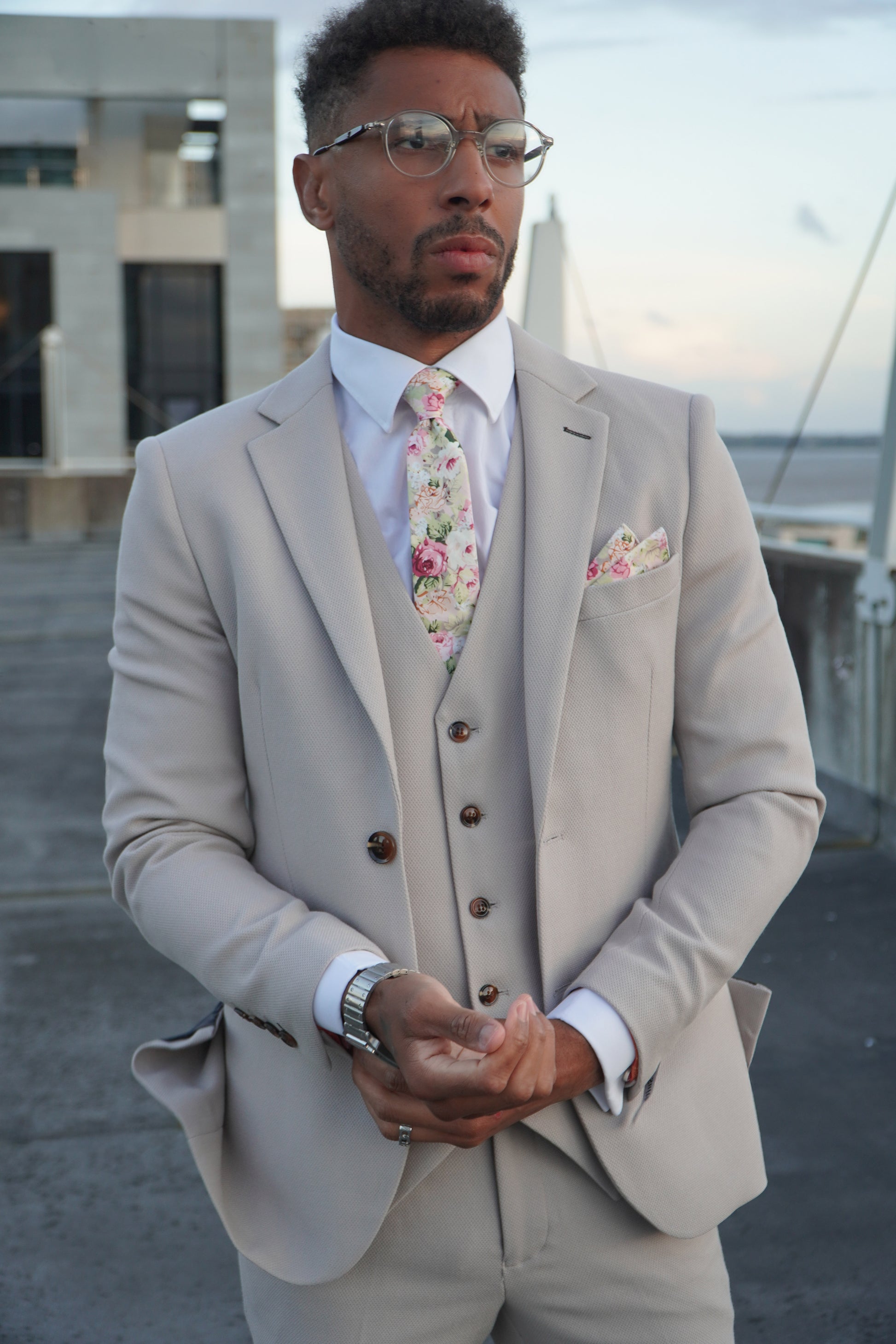 Model wearing the SUITBAE Escobar Stone Three-Piece Suit — tailored beige design with brown buttons, styled with a floral tie and matching pocket square, perfect for weddings and destination events.