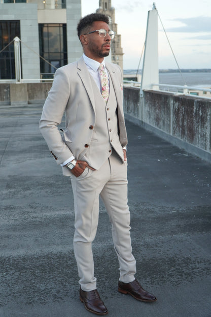 Model wearing the SUITBAE Escobar Stone Three-Piece Suit — beige slim-fit design with brown buttons, paired with a floral tie and dark brown brogues, ideal for weddings and formal summer events.