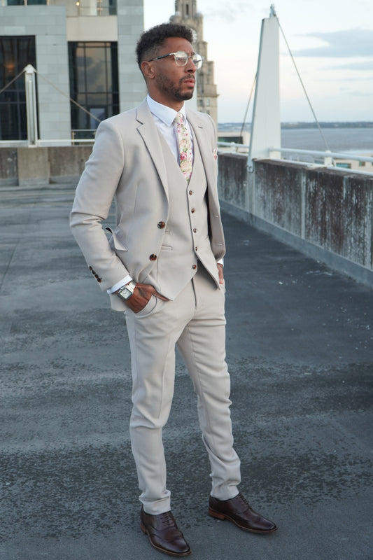 Model wearing the SUITBAE Escobar Stone Three-Piece Suit — beige slim-fit design with brown buttons, paired with a floral tie and dark brown brogues, ideal for weddings and formal summer events.