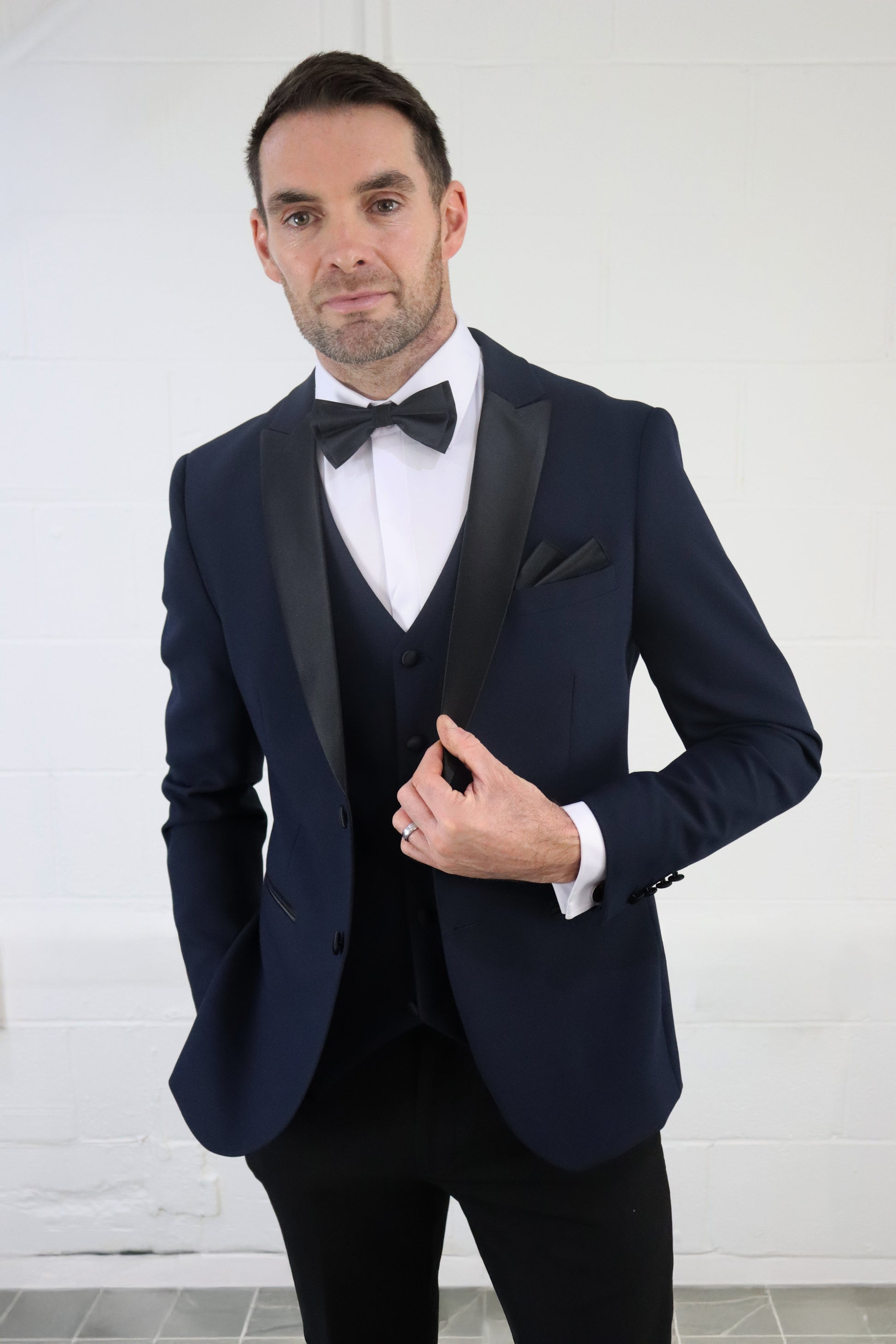 Model wearing the SUITBAE Diablo Navy Three Piece Tuxedo with satin peak lapel, bow tie, and matching waistcoat — perfect for weddings, proms, and black-tie events.