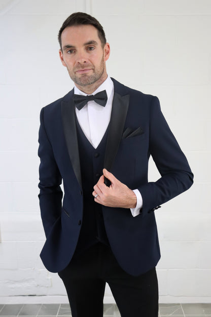 Model wearing the SUITBAE Diablo Navy Three Piece Tuxedo with satin peak lapel, bow tie, and matching waistcoat — perfect for weddings, proms, and black-tie events.
