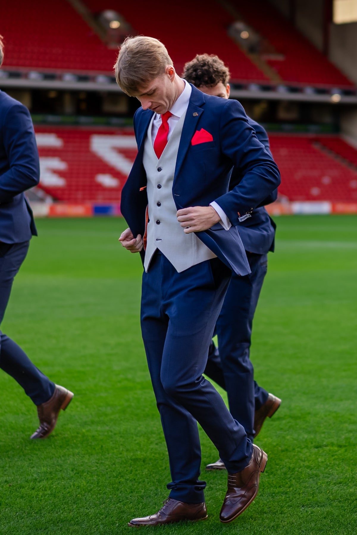 Man wearing the SUITBAE Charles Navy Three-Piece Suit with a red tie and pocket square, walking on the pitch at Barnsley Football Club stadium in brown brogues.