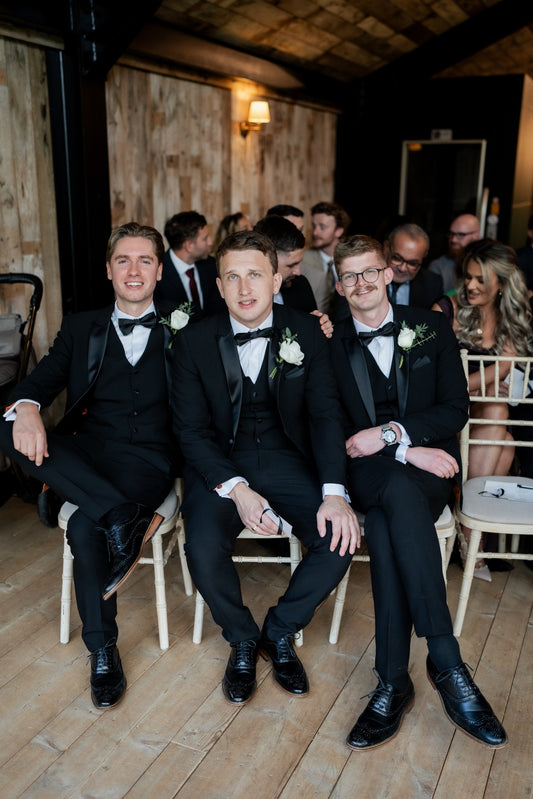 Groomsmen wearing matching SUITBAE Diablo Black Tuxedo Three Piece Suits with satin lapels and bow ties, seated at a wedding ceremony — classic formal menswear style.
