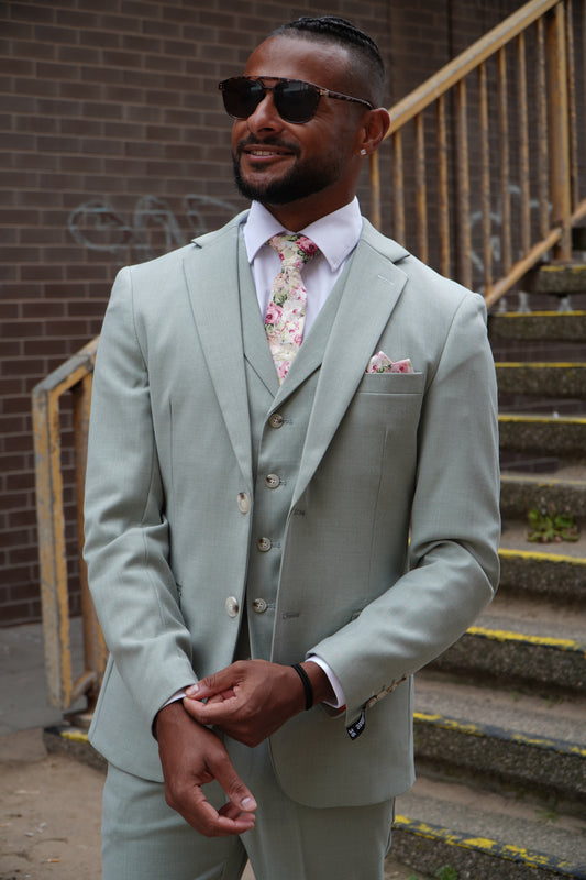 Model wearing the SUITBAE Callum Sage Three-Piece Suit — light sage green slim-fit design with matching waistcoat and trousers, styled with a floral tie and pocket square for weddings and summer events.