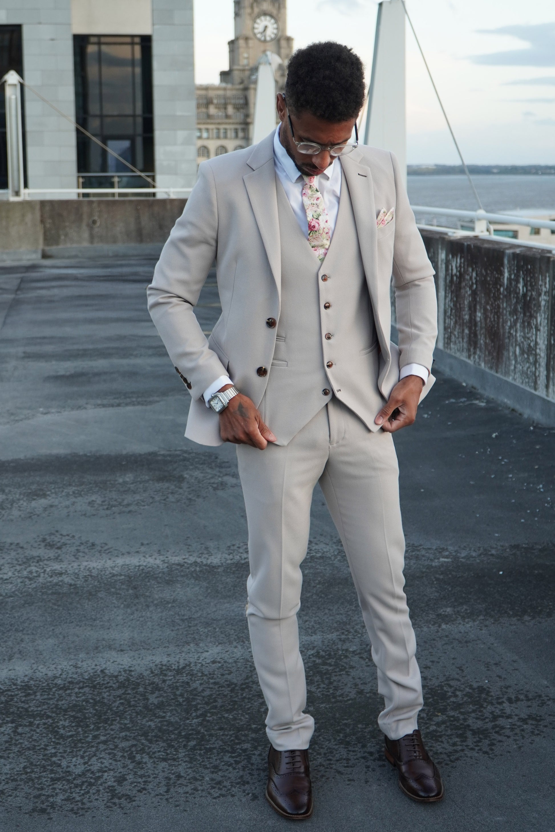Model wearing the SUITBAE Escobar Stone Three-Piece Suit — slim-fit beige design with brown buttons, styled with a floral tie and dark brown brogues, perfect for weddings and summer occasions.
