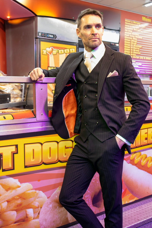 Man wearing a black pinstripe three-piece suit with a white shirt, gold tie, and matching pocket square, standing inside a brightly lit chip shop. The orange inner lining of the jacket is visible, contrasting with the bold yellow and red signage behind him.