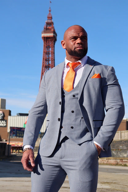 Man wearing the SUITBAE Charles Sky Three-Piece Suit in light blue check with a double-breasted waistcoat, white shirt and tangerine tie, standing by Blackpool Tower — premium men’s wedding, prom and race day suit available in plus sizes.