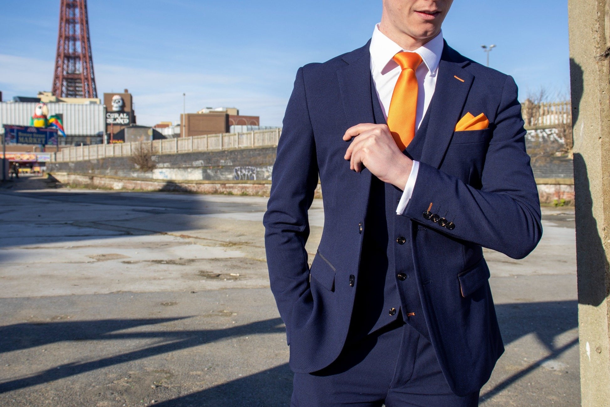 Model wearing the SUITBAE Charles Navy Three-Piece Suit in front of Blackpool Tower. The deep navy fabric features subtle texture and contrast tangerine stitching, paired with an orange tie and pocket square for a bold SUITBAE signature look. Available in plus sizes up to 58" chest and 52" waist.