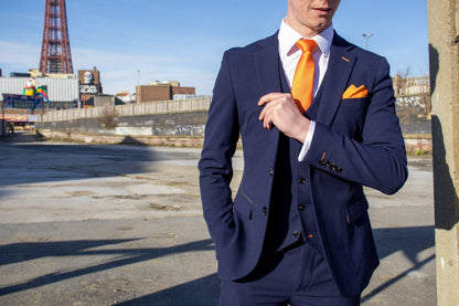 Model wearing the SUITBAE Charles Navy Three-Piece Suit in front of Blackpool Tower. The deep navy fabric features subtle texture and contrast tangerine stitching, paired with an orange tie and pocket square for a bold SUITBAE signature look. Available in plus sizes up to 58" chest and 52" waist.