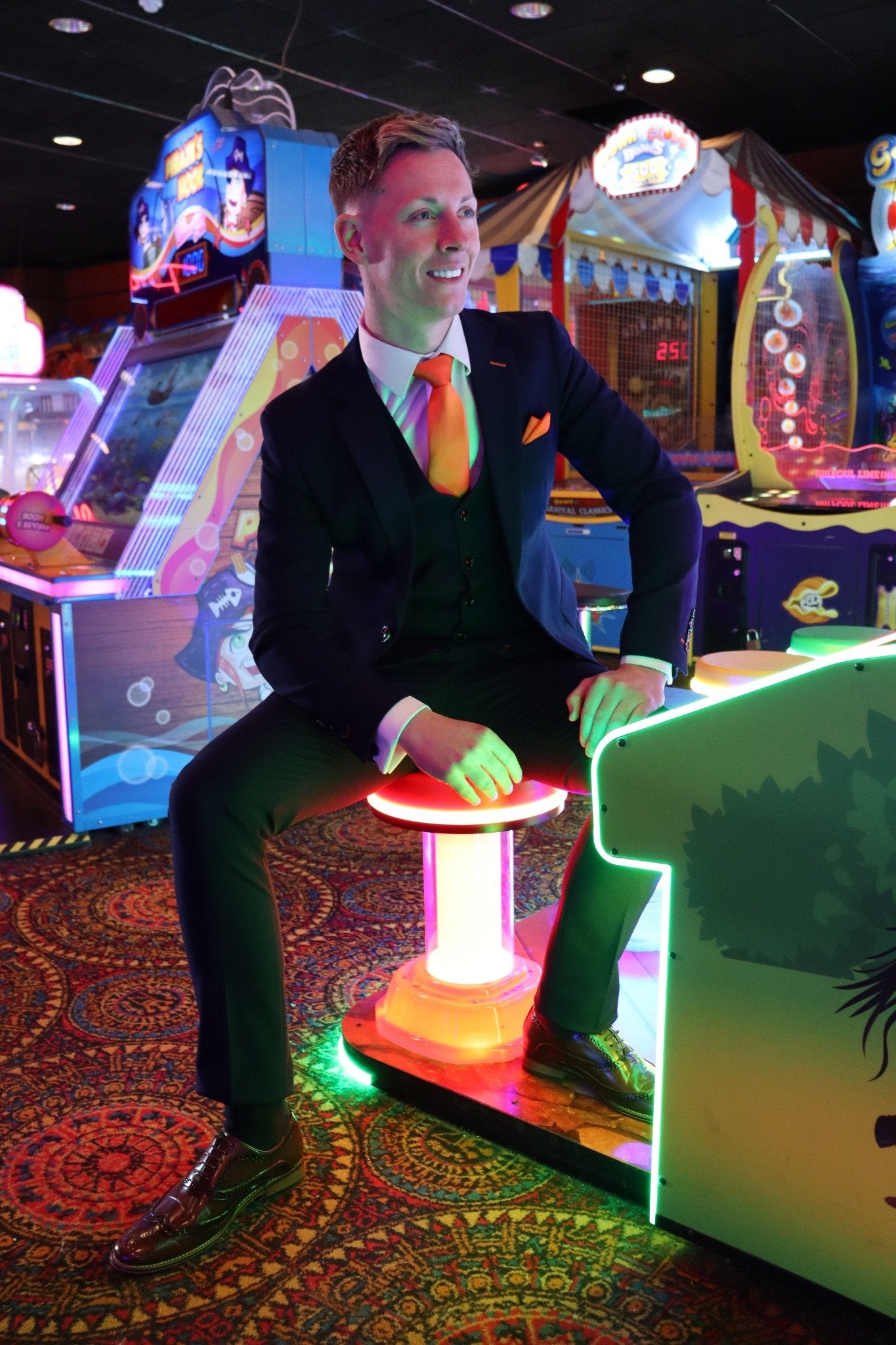Model sitting inside a Blackpool arcade wearing the SUITBAE Charles Navy Three-Piece Suit. The deep navy fabric contrasts with the glowing arcade lights, styled with a bright orange tie and pocket square for a bold, confident look. Available in plus sizes.