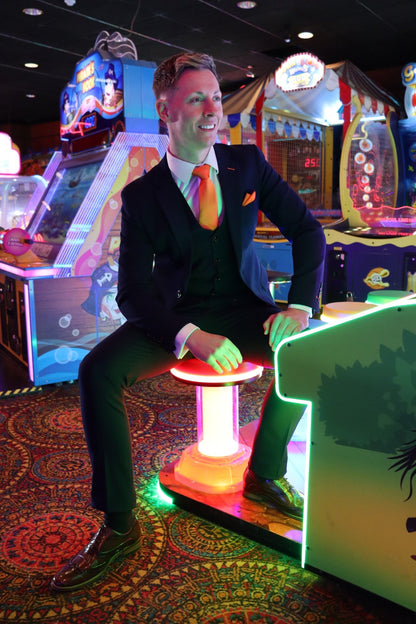 Model sitting inside a Blackpool arcade wearing the SUITBAE Charles Navy Three-Piece Suit. The deep navy fabric contrasts with the glowing arcade lights, styled with a bright orange tie and pocket square for a bold, confident look. Available in plus sizes.