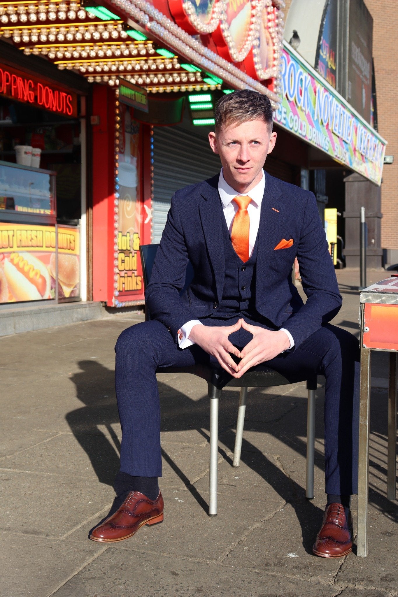 Model seated in the SUITBAE Charles Navy Three-Piece Suit outside a seaside shop. The rich navy fabric is paired with a crisp white shirt, tangerine tie, and matching pocket square — capturing the Blackpool heritage and signature SUITBAE flair. Available in plus sizes.