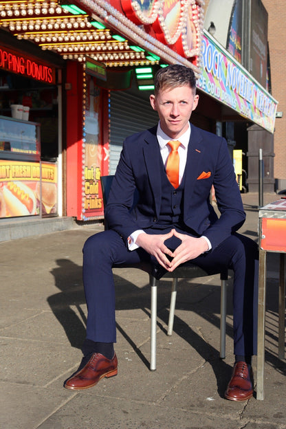 Model seated in the SUITBAE Charles Navy Three-Piece Suit outside a seaside shop. The rich navy fabric is paired with a crisp white shirt, tangerine tie, and matching pocket square — capturing the Blackpool heritage and signature SUITBAE flair. Available in plus sizes.