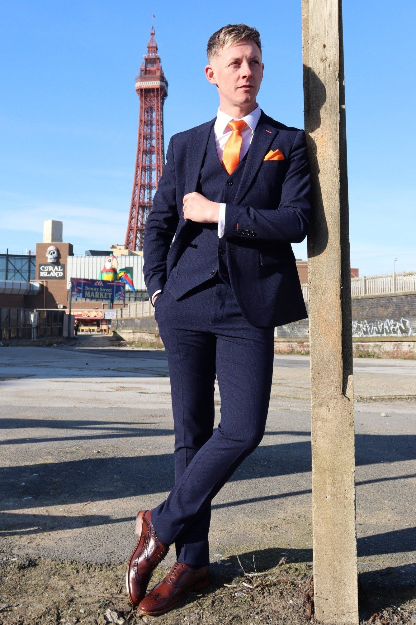 Model wearing the SUITBAE Charles Navy Three-Piece Suit in front of Blackpool Tower. The suit’s rich navy tone is paired with an orange tie and pocket square, showcasing SUITBAE’s tangerine signature style. Perfect for weddings, races, or formal events. Available in plus sizes.