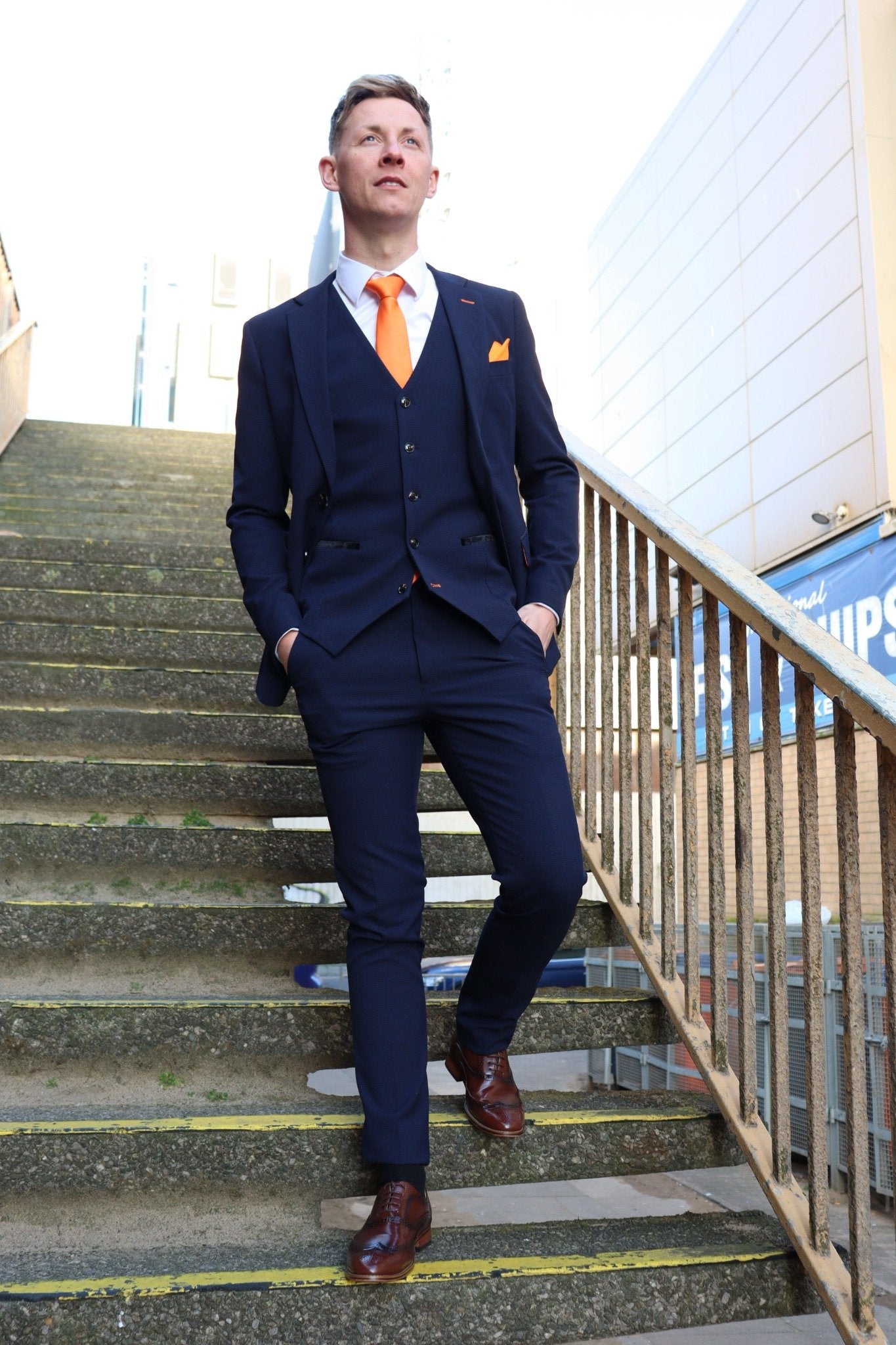 Model walking down steps wearing the SUITBAE Charles Navy Three-Piece Suit paired with an orange tie and pocket square. The navy textured fabric contrasts sharply with the tangerine accessories, reflecting SUITBAE’s Blackpool-inspired signature style. Available in plus sizes.