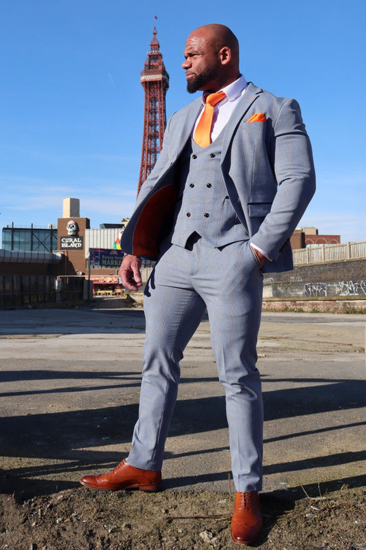 Man wearing the SUITBAE Charles Sky Three-Piece Suit in light blue check, featuring a double-breasted waistcoat and tangerine lining, styled with orange tie and pocket square on Blackpool Promenade — tailored fit ideal for weddings, proms and races, available in plus sizes.