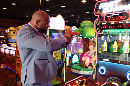 Man wearing the SUITBAE Charles Sky Three-Piece Suit in a light blue check pattern inside a colourful arcade, styled with a tangerine tie and pocket square — a tailored slim-fit men’s suit ideal for weddings, races and proms, available in plus sizes.