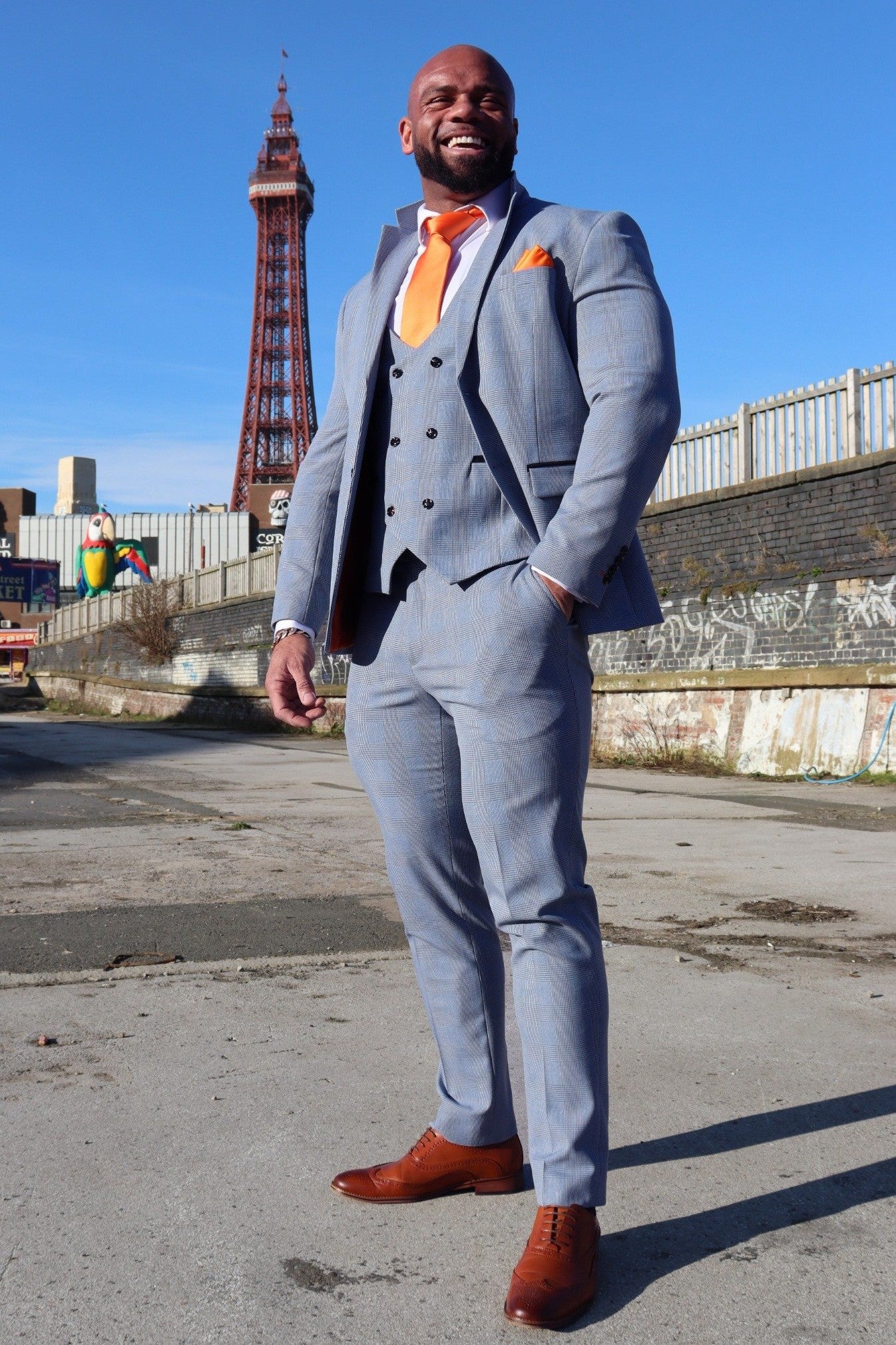 Model wearing the SUITBAE Charles Sky Three-Piece Suit in light blue check on Blackpool Promenade with the iconic tower behind, styled with tan brogues and a tangerine tie — a slim-fit wedding, prom, or race day suit available in plus sizes.