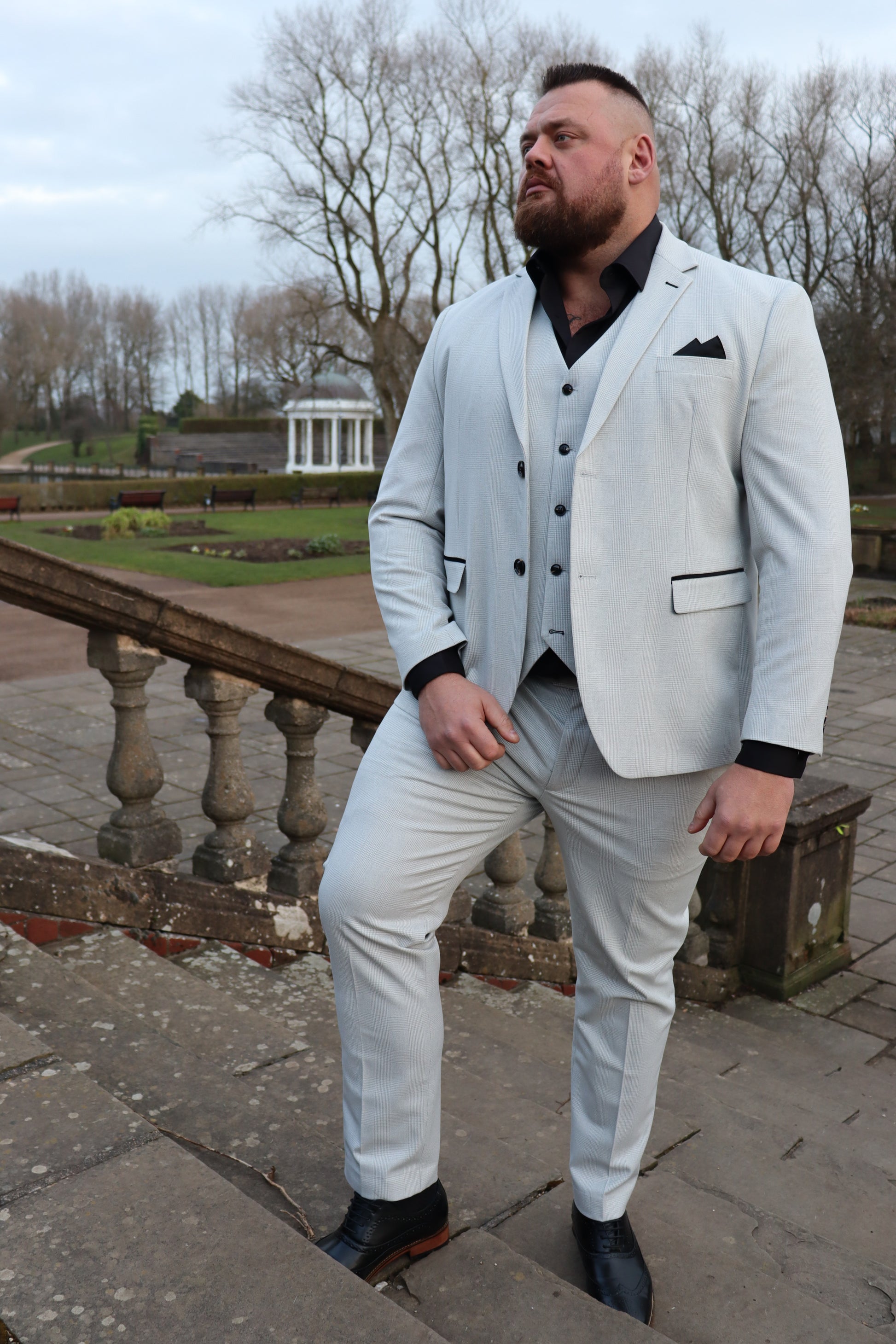 Model wearing the SUITBAE Charles Stone Three-Piece Suit with a black shirt and black brogues. The light stone fabric with subtle patterning and black contrast trim delivers a clean, structured finish. Tailored for comfort and style, available in plus sizes for weddings, proms, and formal occasions.