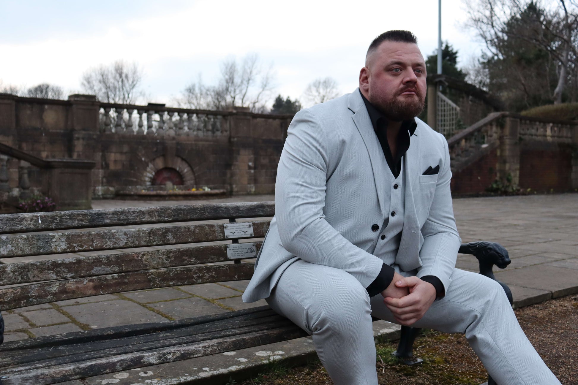 Model seated on a park bench wearing the SUITBAE Charles Stone Three-Piece Suit, styled with a black shirt and pocket square. The light stone fabric and black contrast details create a sharp, structured look. Available in plus sizes, designed for comfort and standout presence at weddings, proms, and formal occasions.