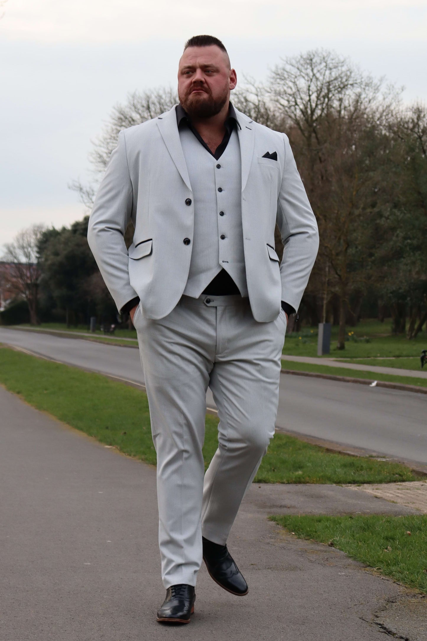 Model walking confidently in the SUITBAE Charles Stone Three-Piece Suit, featuring a light stone textured fabric with black contrast trim and buttons. Styled with a black shirt and pocket square, this modern formal look offers structure and stretch comfort. Available in plus sizes, perfect for weddings, races, and special occasions.