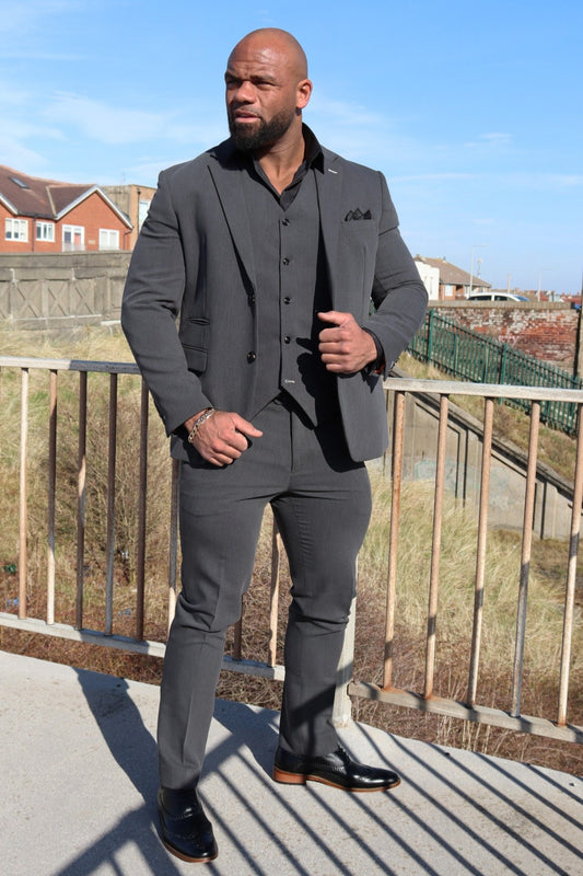 Man wearing a charcoal grey three-piece suit with a black shirt and black pocket square, standing outdoors on a sunny day. The fitted suit is paired with polished black shoes featuring tan soles, highlighting a modern and confident look.