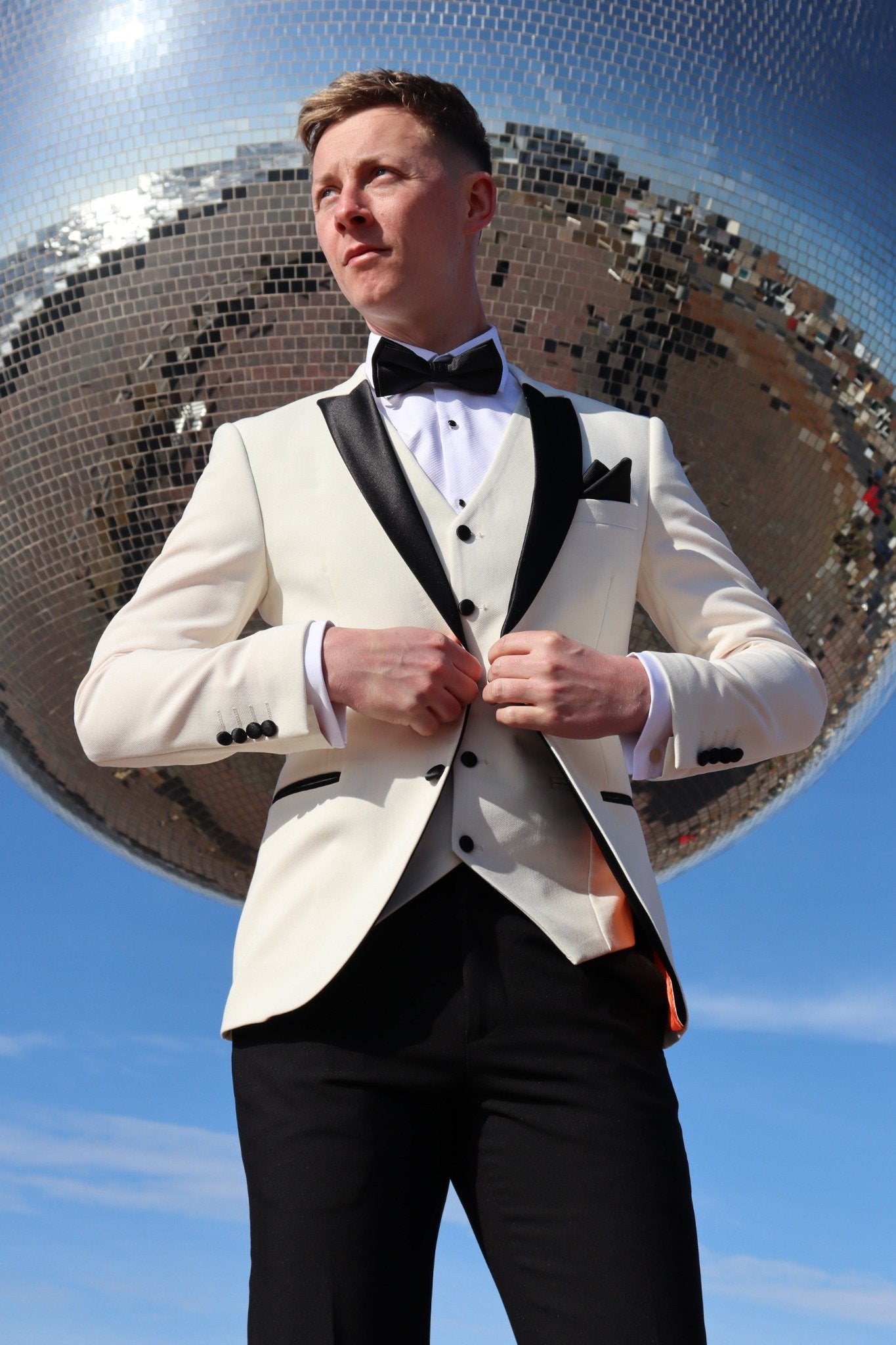 Model wearing the SUITBAE Diablo Ivory Tuxedo in front of Blackpool’s Mirror Ball, featuring black satin lapels and a matching waistcoat — luxury men’s three-piece tuxedo perfect for weddings, black-tie events and proms.