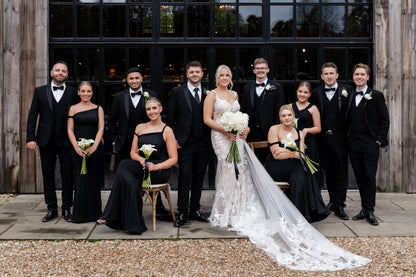 Groom and groomsmen wearing SUITBAE Diablo Black Tuxedo Three Piece Suits with satin lapels, styled with white shirts and bow ties beside bridesmaids in black dresses — perfect black-tie wedding look.