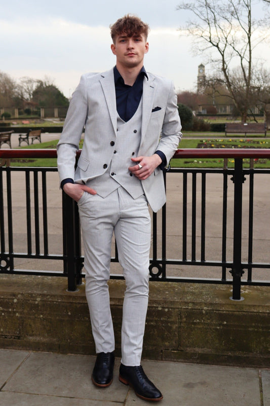 Man wearing a light grey three-piece suit with a dark navy shirt and pocket square, paired with black formal shoes, standing by a park railing — part of the SUITBAE menswear collection.