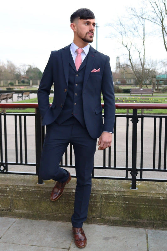 A man wearing the Lawrence navy three piece suit with brown buttons, a white shirt, and a mauve tie and pocket square leaning on a metal railing.