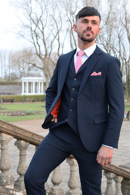 Man wearing a navy three-piece suit with a pink tie and pocket square, posing on stone steps in a park with a white pavilion in the background — part of the SUITBAE formalwear collection.