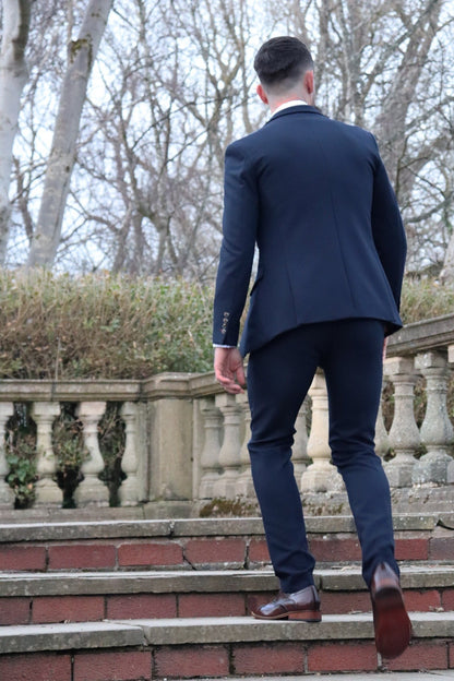 Man walking up stone steps wearing a fitted navy suit and brown brogue shoes, photographed from behind in an outdoor park setting — part of the SUITBAE formalwear collection.