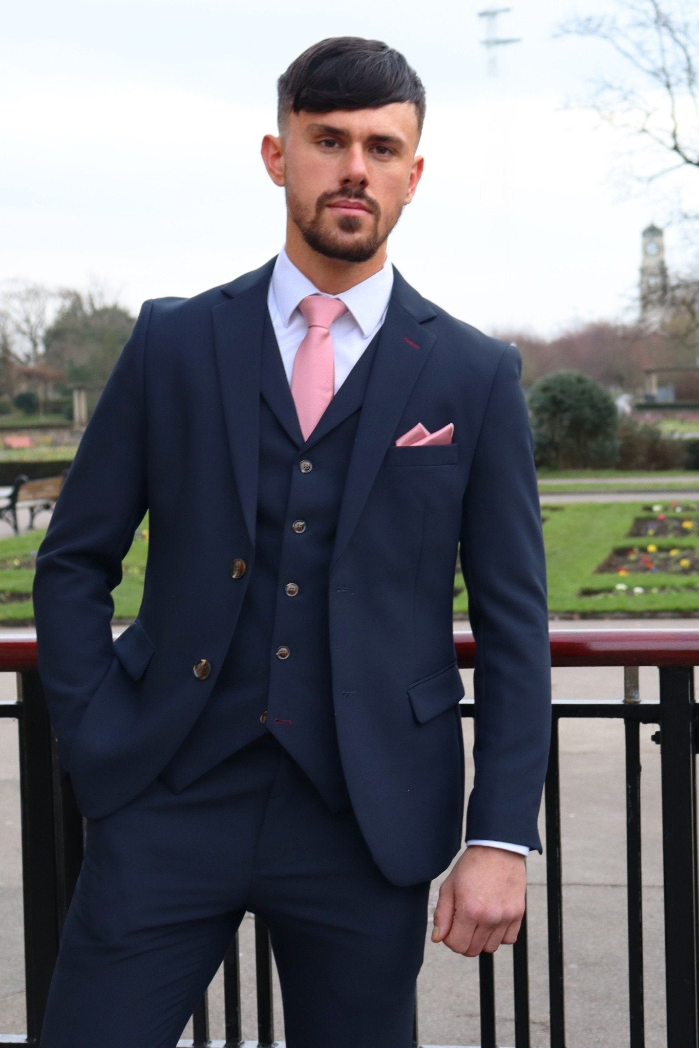 Man wearing a navy three-piece suit with a pink tie and pocket square, standing by a park railing with gardens in the background — part of the SUITBAE formalwear collection.