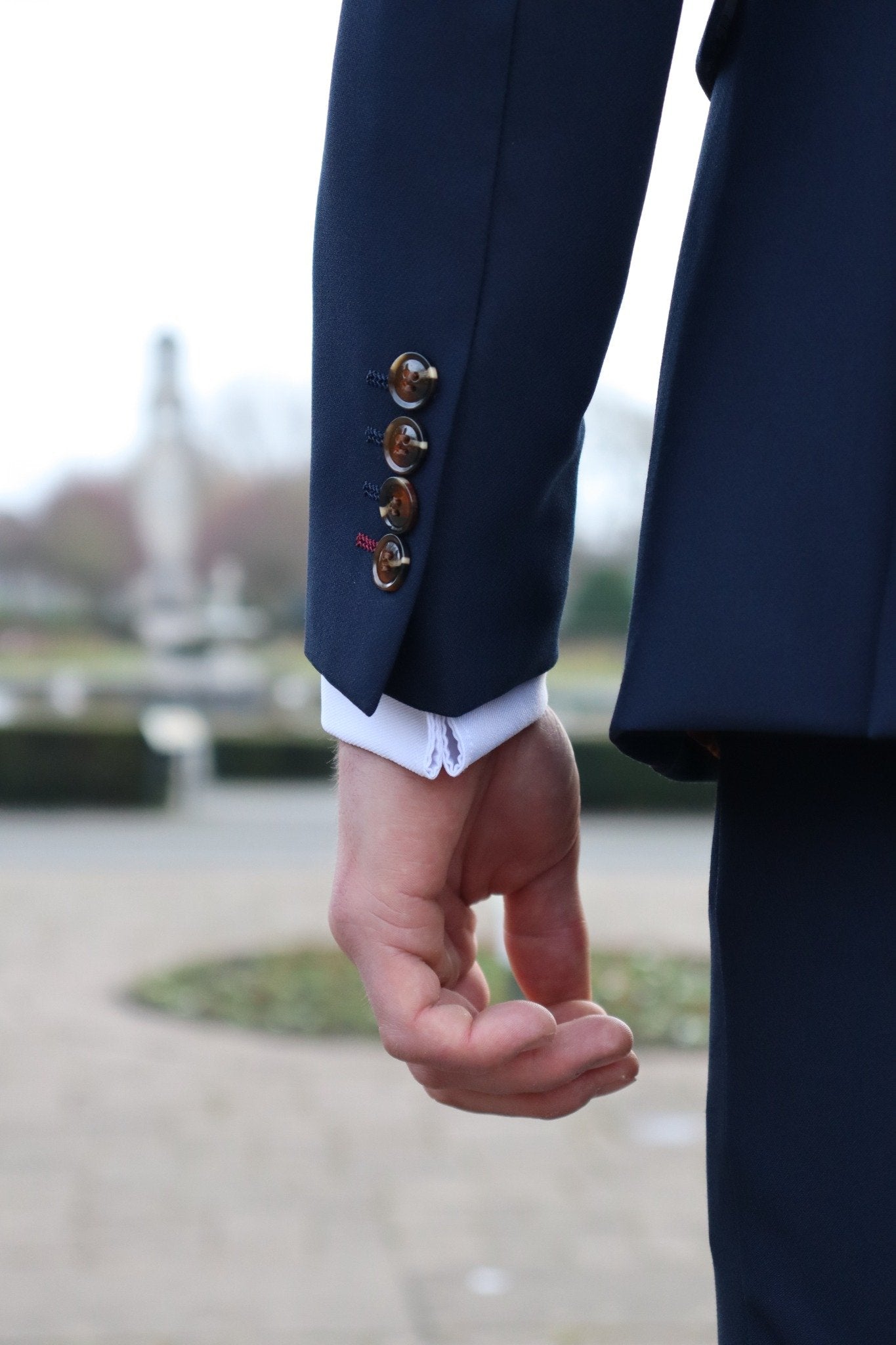 Close-up of a man’s arm wearing a navy suit jacket with brown buttons and visible white shirt cuff, showing the sleeve detail and stitching from the SUITBAE collection.